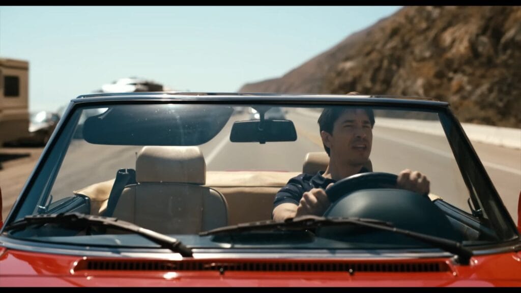 Justin Long as AJ driving a red convertible down a sunny coastal highway in Barbarian.