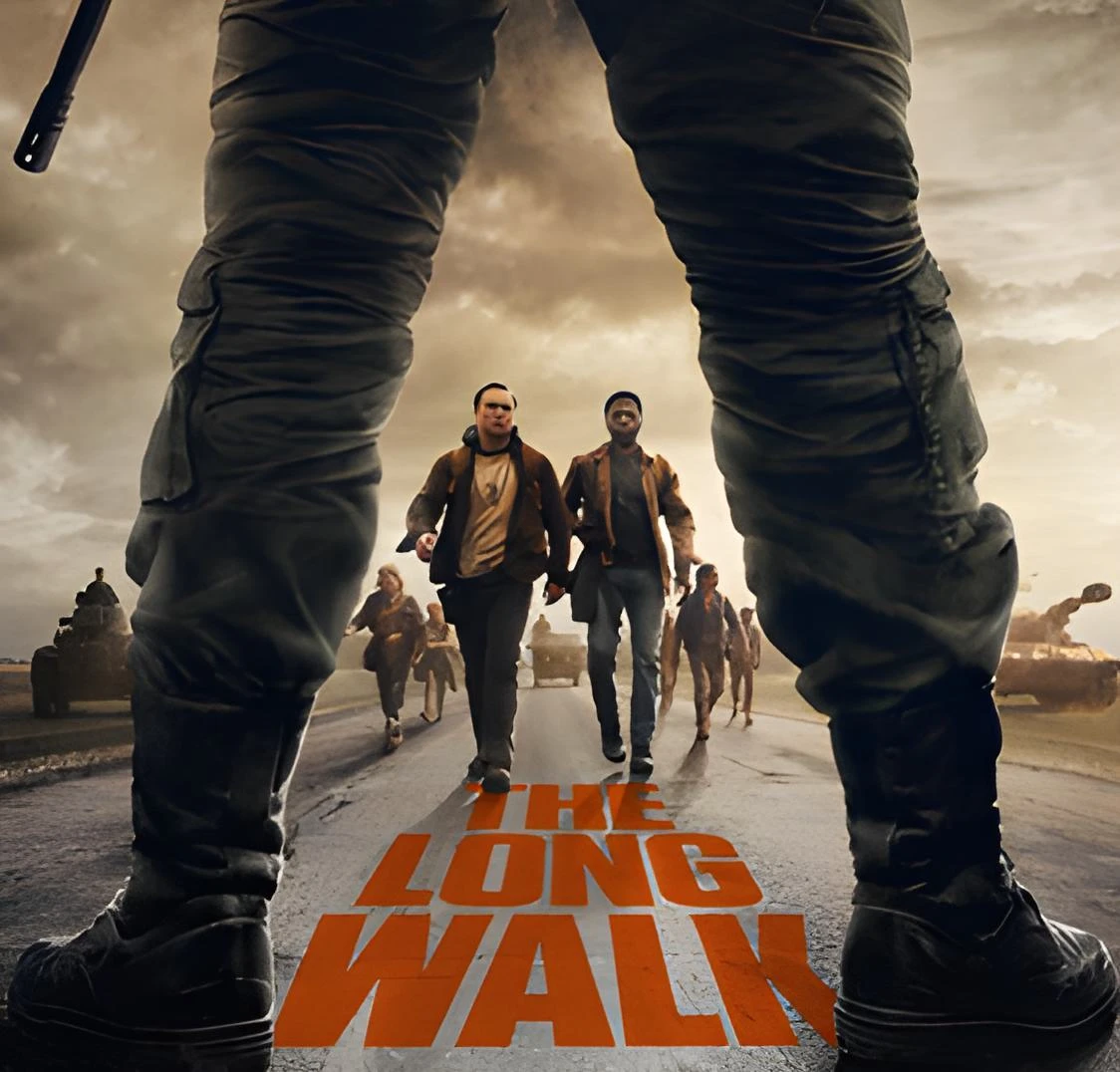 A low-angle perspective shot looking through the legs of a soldier in dark combat trousers and boots. In the distance, two men walk down a paved road toward the viewer, followed by a group of weary figures and flanked by military tanks. Large orange text painted on the road surface reads "THE LONG WALK."