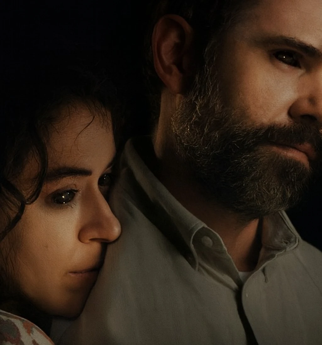 Close-up of Tatiana Maslany as Liz resting her head on the shoulder of Rossif Sutherland as Malcolm in the horror movie Keeper (2025), with dark, moody lighting.