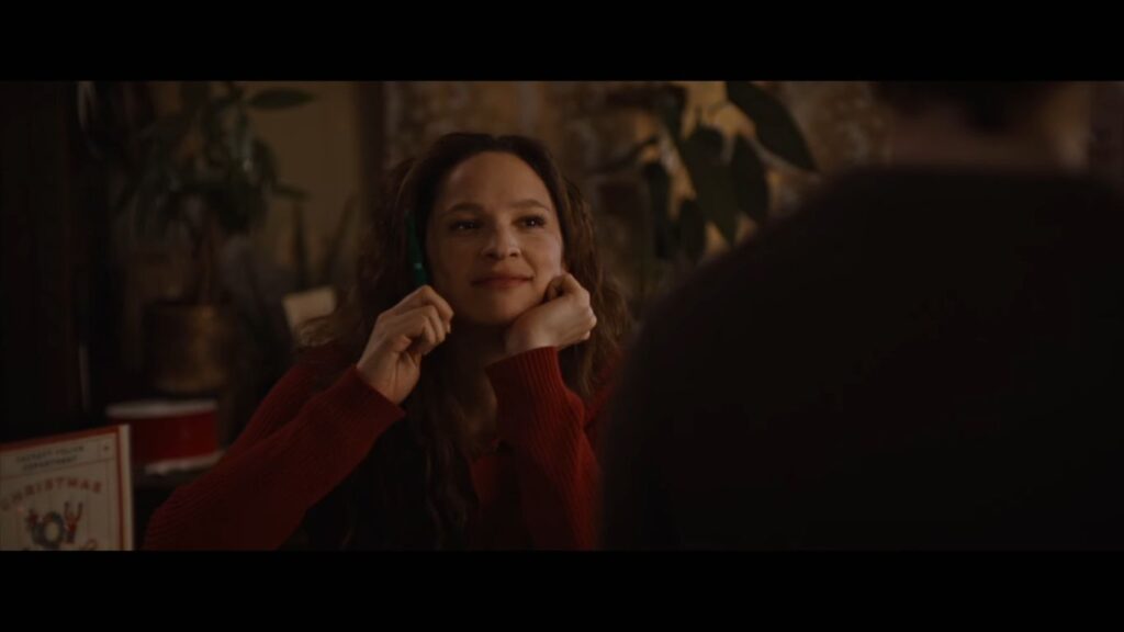 Pamela (Ruby Modine) smiles softly in Silent Night, Deadly Night (2025), resting her chin on her hands with a 'Christmas Toy' sign visible.
