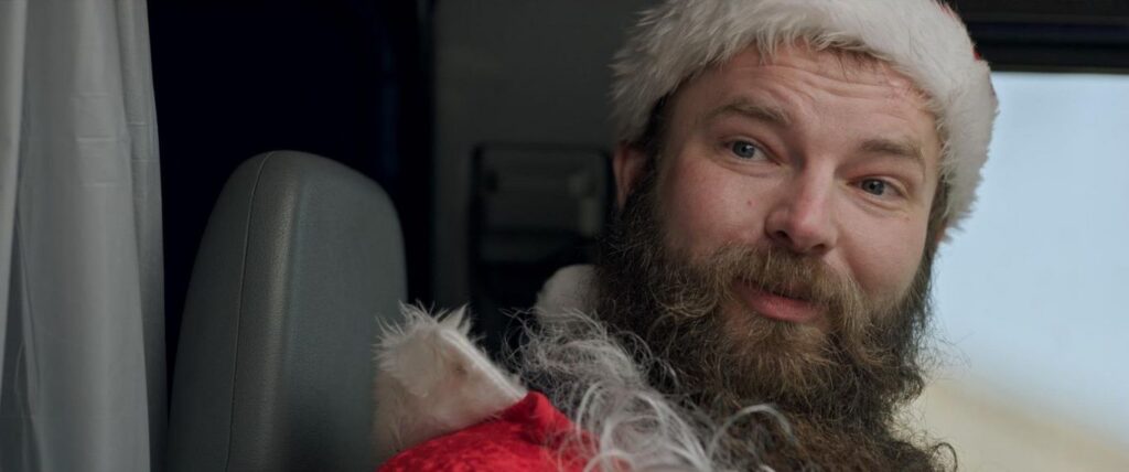 A close-up of a bearded man wearing a Santa hat looking into the camera in The Christmas Spirit (2023), capturing the bizarre energy of low budget Christmas horror movies.
