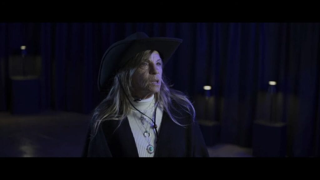 Laurie Felix Bass as Dolly Broadbent, wearing a black cowboy hat and shawl, looking off-camera with a serious expression in a dimly lit interview setting.