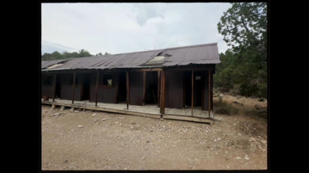 A dilapidated wooden shack in Horror in the High Desert 4: Majesty with a rusty, collapsing metal roof sitting in a dry, rural landscape, presented in a 4:3 aspect ratio.
