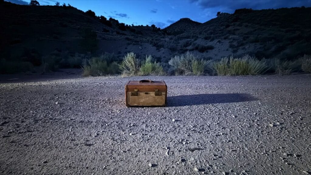 A worn brown suitcase from Horror in the High Desert 4: Majesty sits alone on the gravel ground of the high desert at twilight, illuminated by a spotlight against a backdrop of dark hills.