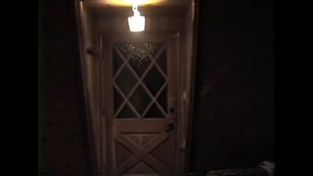 A grainy, found-footage style shot of a wooden door with a diamond-patterned window, illuminated by a single overhead light in the pitch darkness.