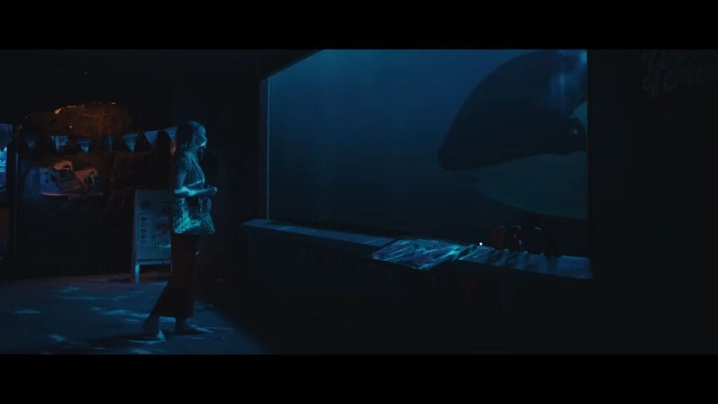 A character staring into a large, blue-lit aquarium tank where the silhouette of a massive orca is visible.