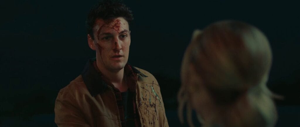 A medium shot of a young man with dark hair, looking distressed with blood on his forehead and splattered across his tan jacket, facing a woman whose back is turned to the camera.