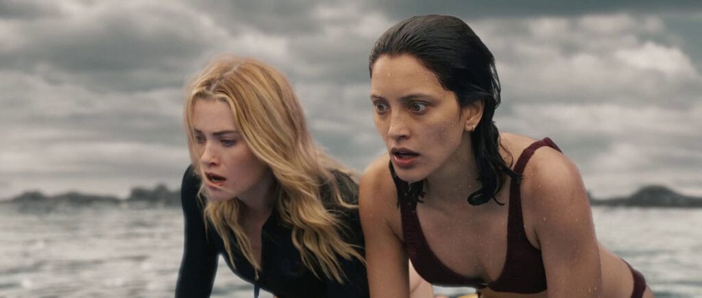 Close-up of two young women, one blonde and one brunette, appearing wet and looking terrified while in the ocean against a backdrop of a grey, overcast sky.