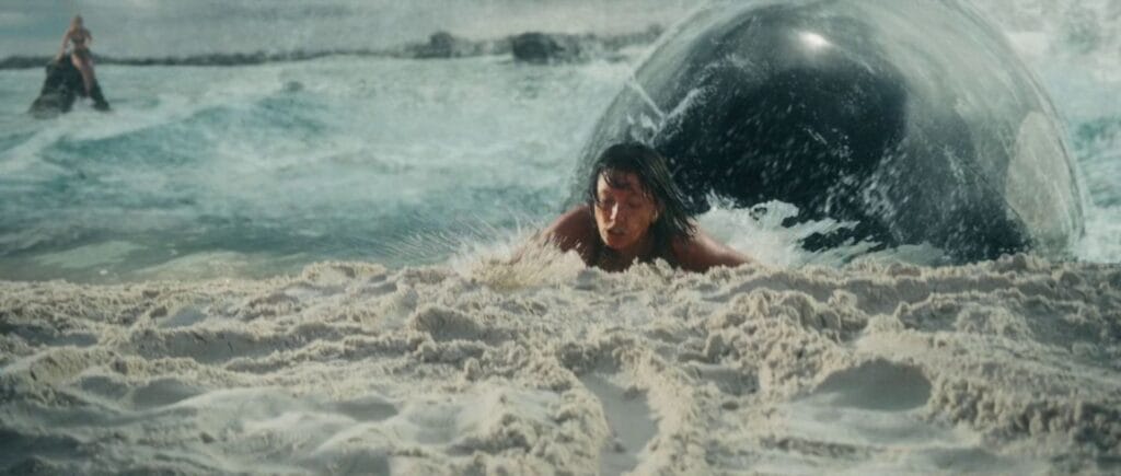 A dark-haired woman, Trish, frantically crawls up a sandy beach as a massive black whale lunges out of the water behind her, while another woman stands near the water's edge in the background.