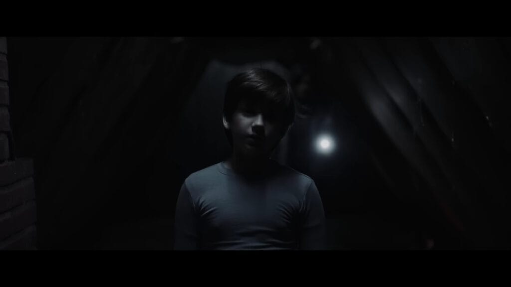 A young boy standing in a dark, eerie attic during the climax of The Confession (2025).