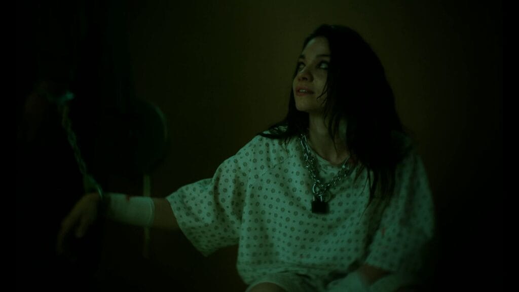 Paloma sits in a dark room wearing a hospital gown and heavy metal chains around her neck and wrists, reflecting the psychological thriller elements of the Twisted ending.
