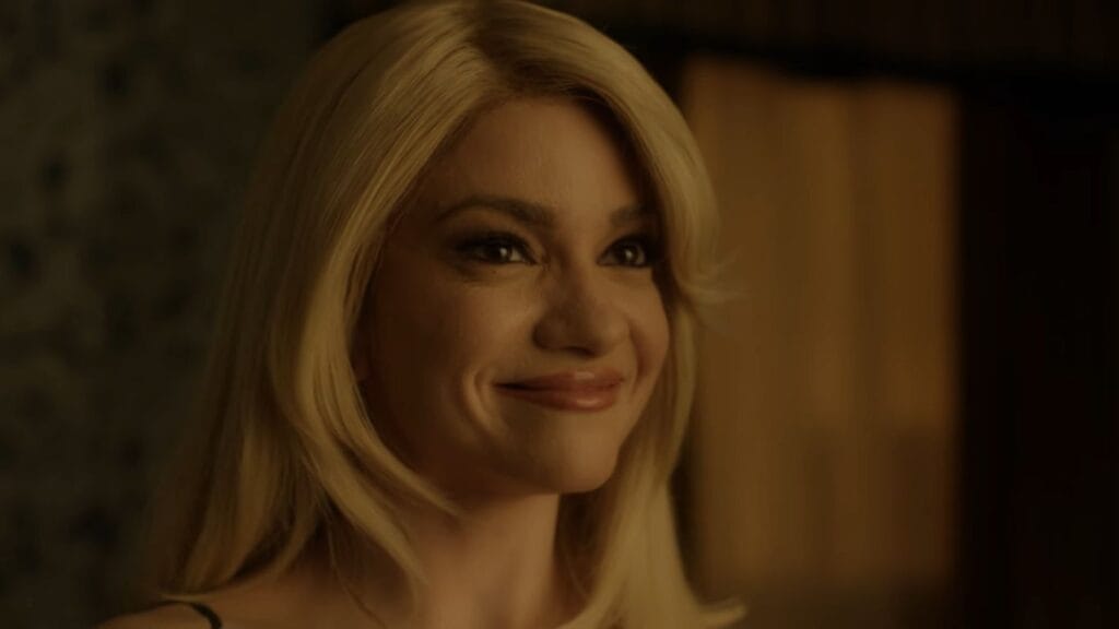 Close-up of Paloma (Lauren LaVera) with blonde hair, wearing a faint, unsettling smile in a warm-toned indoor setting.