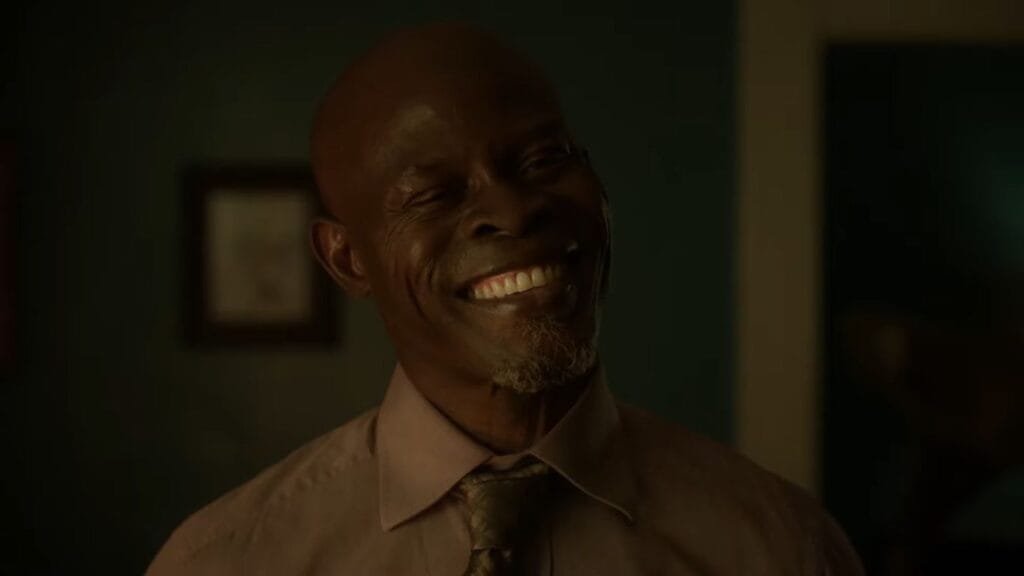 Dr. Kezian (Djimon Hounsou) in a dress shirt and tie, smiling widely with an expression that appears menacing or unhinged.