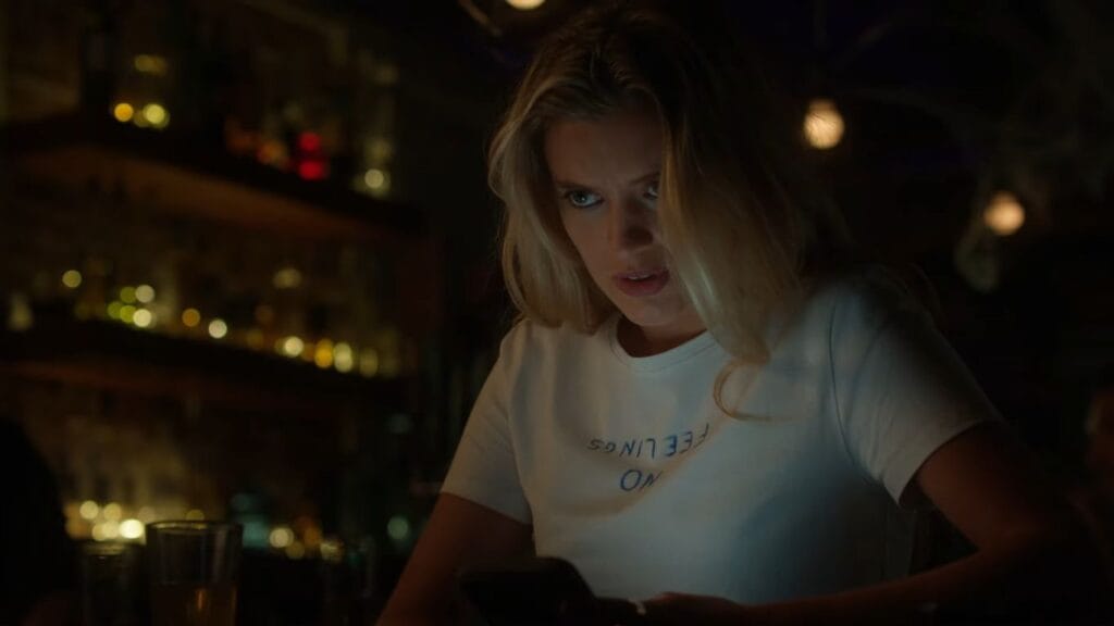 Smith (Mia Healey) looking intensely at her phone in a dimly lit bar, wearing a white t-shirt that reads 'NO FEELINGS ON'.