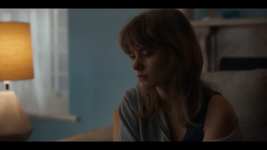 A cinematic still from "Diabolic" showing a young woman with bangs and light brown hair sitting in a dimly lit room. She has a somber, contemplative expression as she looks downward. A warm, glowing lamp is visible in the blurred background.