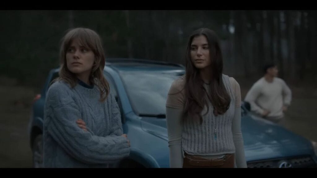 Two women standing outside in a wooded area next to a blue SUV. The woman in the foreground wears a thick, blue cable-knit sweater and looks off to the side with a concerned expression. A man stands in the distant background.