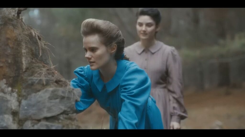 A scene from "Diabolic" featuring two women in a forest. The woman in the foreground wears a bright blue, vintage-style dress with puffed sleeves and is leaning over a mossy stone ruin. Another woman in a muted brown dress stands behind her.