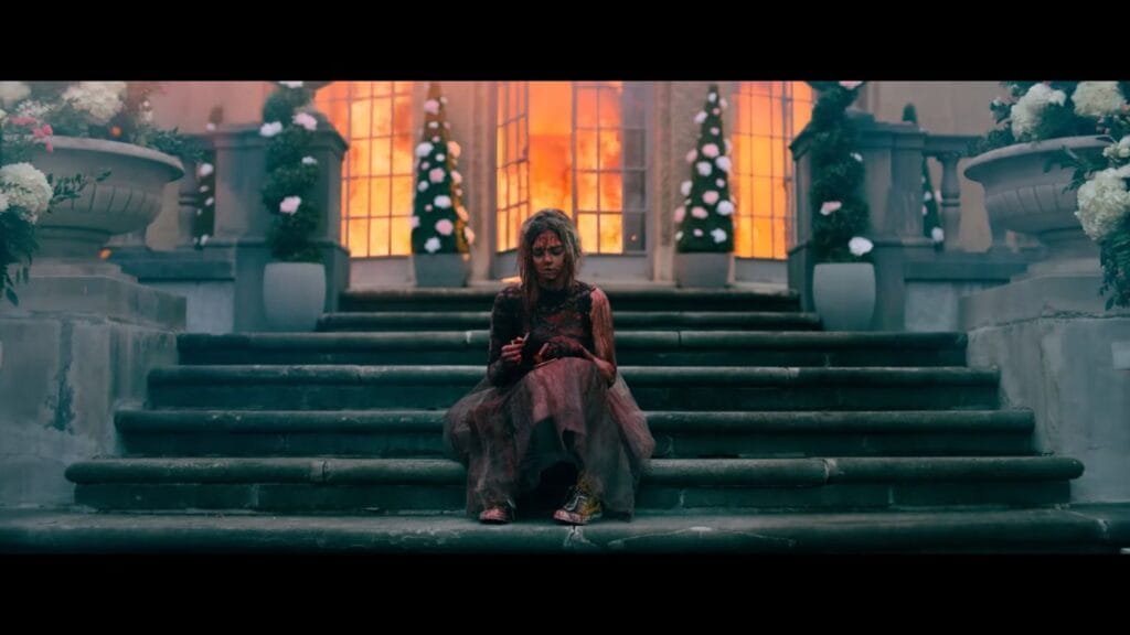 A blood-splattered Grace (Samara Weaving) sits on the stone steps of a mansion as a fire rages in the background in a scene from Ready or Not 2: Here I Come (2026).