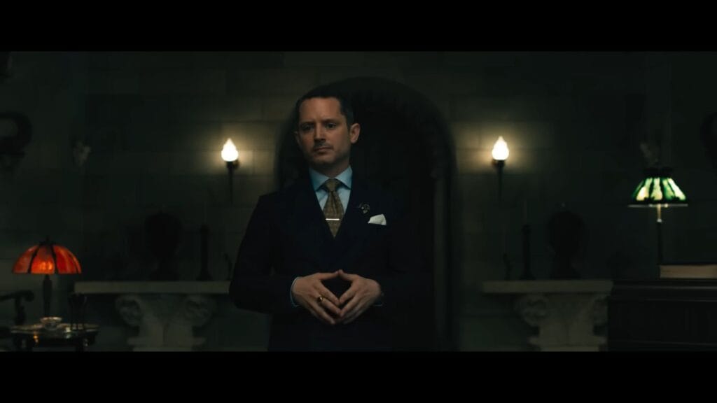 The Lawyer (Elijah Wood) stands in a dark, gothic room wearing a navy suit and gold tie, looking menacing in the 2026 horror film Ready or Not 2.