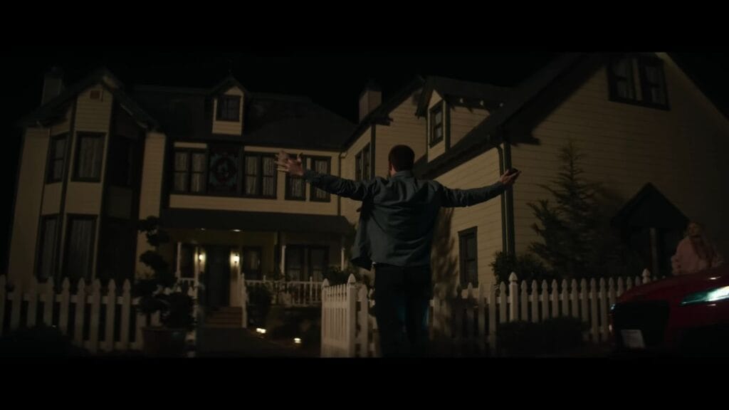 A man stands with his arms outstretched in front of a large, two-story suburban house at night. A red car is partially visible in the foreground, and the scene is lit by warm exterior house lights against a dark sky.