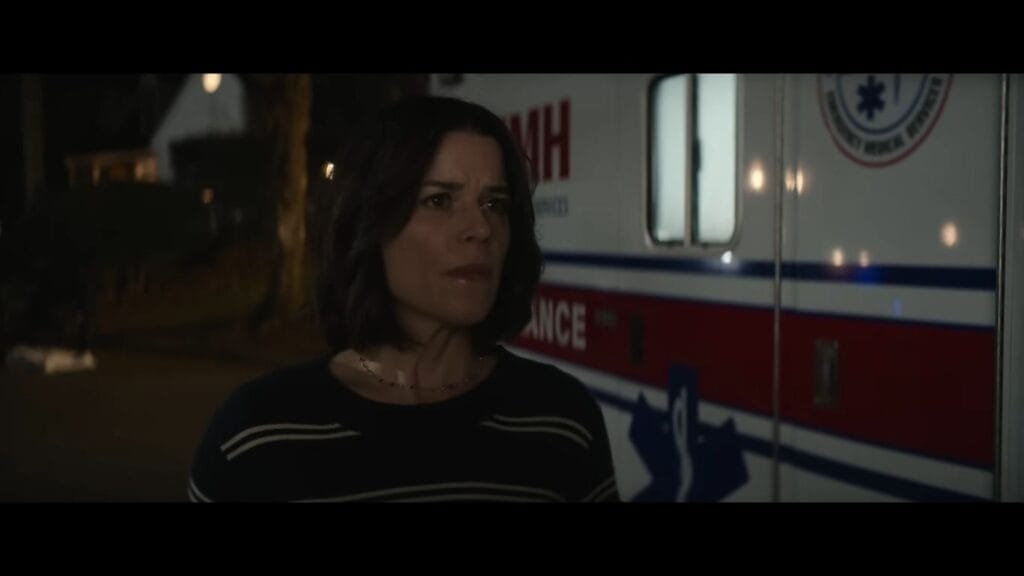 Sidney Prescott (Neve Campbell) looking concerned and alert while standing in front of an ambulance at night. She is wearing a dark, striped sweater, and the red and blue emergency vehicle markings are visible behind her.