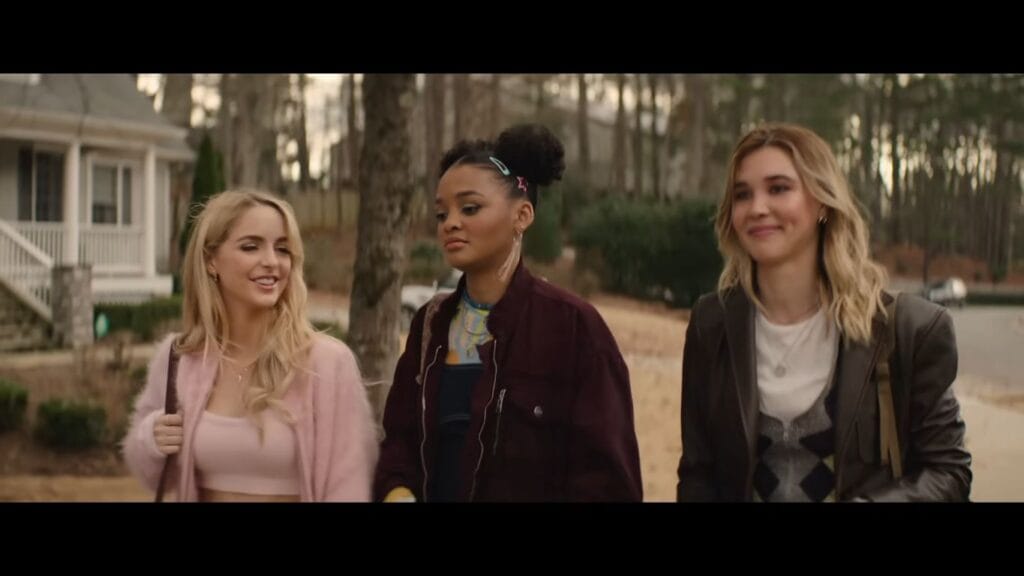 Three young women walking together outdoors during the day. The woman on the left wears a pink fuzzy cardigan, the center woman has her hair in space buns with a maroon jacket, and the woman on the right wears a black leather jacket. They appear to be in a residential neighborhood.