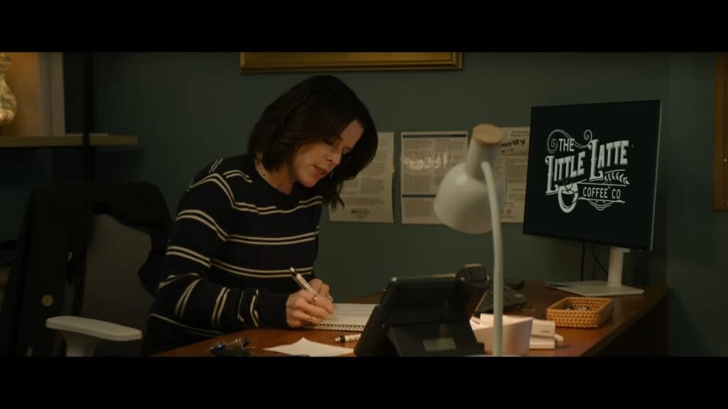 Sidney Prescott (Neve Campbell) sitting at a desk in an office, focused on writing in a notebook. A computer monitor to her right displays a logo for "The Little Latte Coffee Co." against a dark green wall.