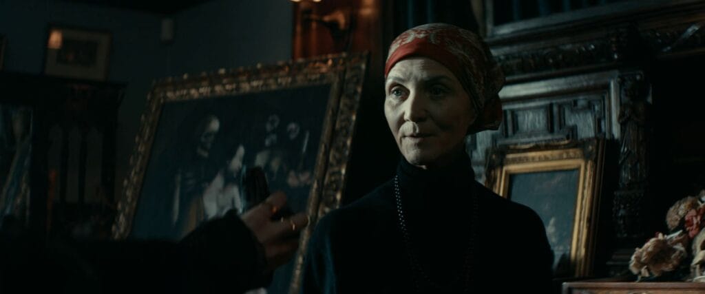 Ivy, an older woman wearing a red patterned headscarf and black turtleneck, looks on tensely in a dimly lit room in the horror movie Whistle (2025).