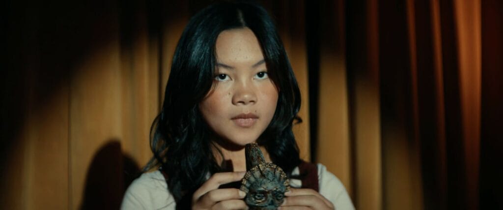 Asha, a young girl, holds the cursed ancient skull whistle with both hands against a backdrop of gold curtains in the horror film Whistle.