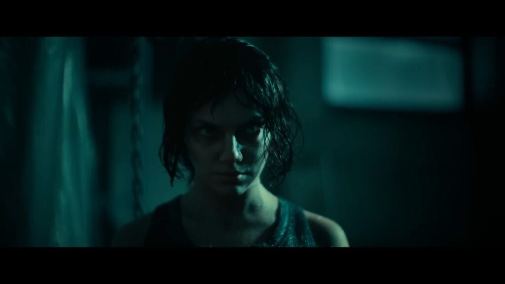 Chrys (Dafne Keen) stares intensely with a haunting expression in a dark, teal-lit room in a scene from the horror movie Whistle (2025).