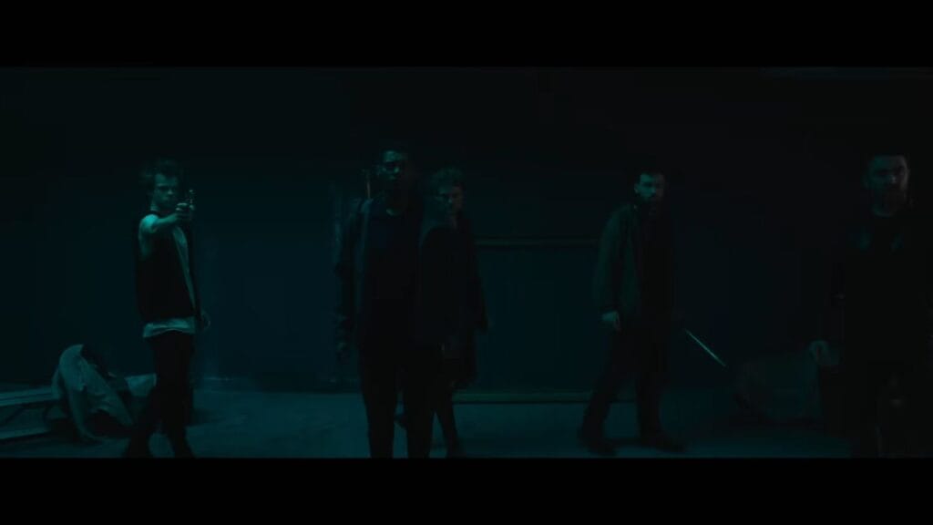 A group of survivors stands in a dark, teal-lit room, with one man pointing a gun into the shadows in the 2026 horror film Do Not Enter.