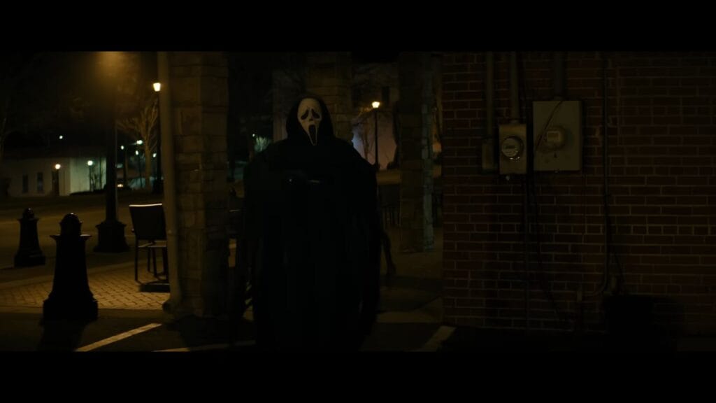 Ghostface stands in the dark under an outdoor brick archway, a classic horror visual from the 2026 movie Scream 7.