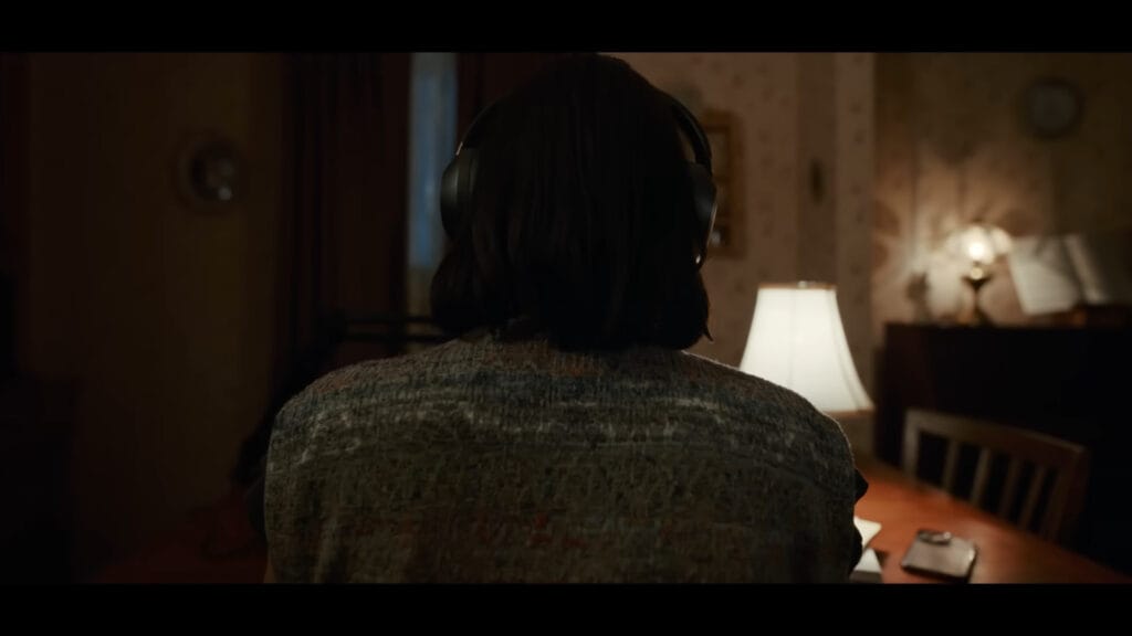 Nina Kiri as Evy seen from behind, wearing headphones at a desk in a dimly lit room.