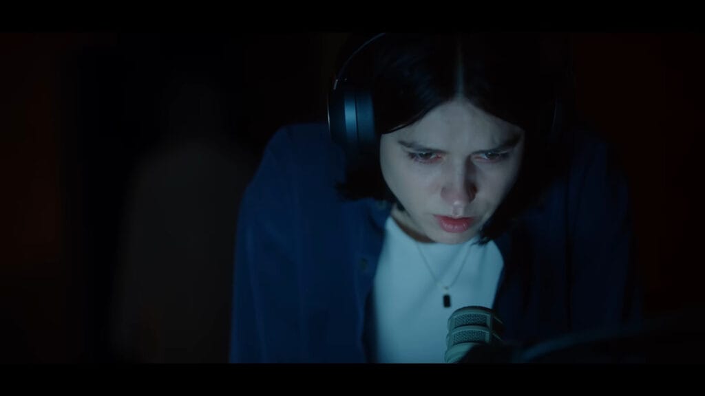 Evy (Nina Kiri) hunched over a microphone under moody blue lighting, looking distressed.