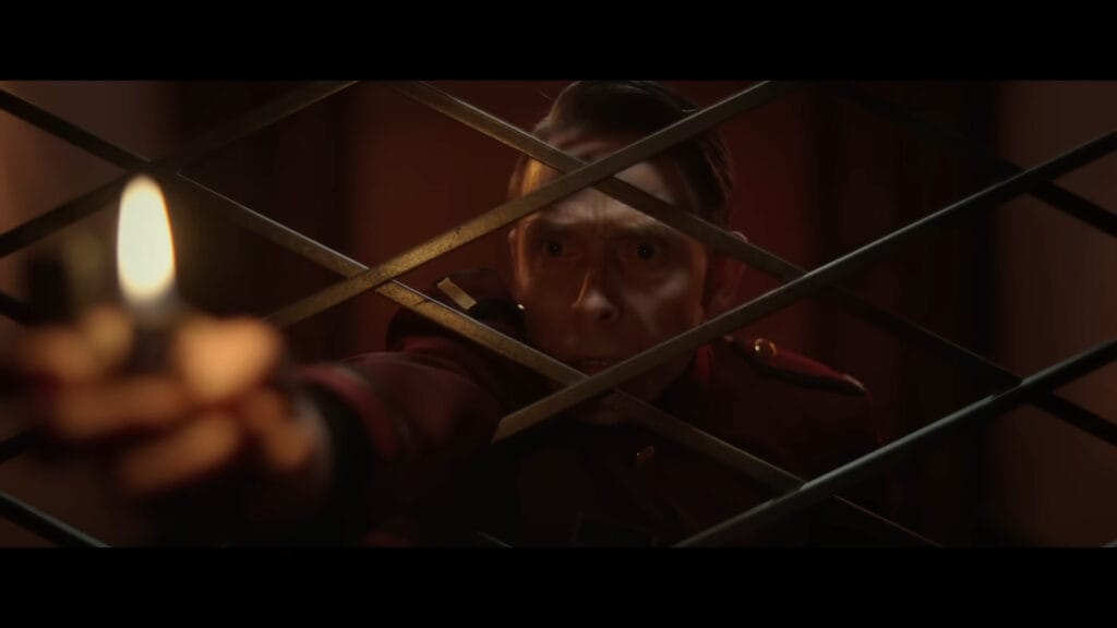 A man in a red hotel uniform looking nervously through a metal gate while holding a lit lighter.