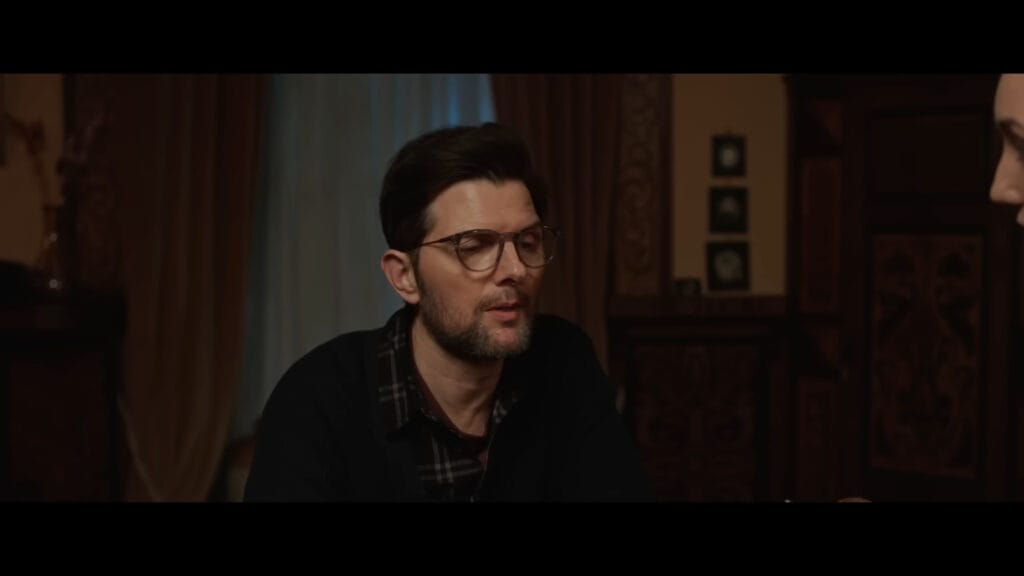 Adam Scott wearing glasses and a dark cardigan sitting in a dimly lit hotel room.