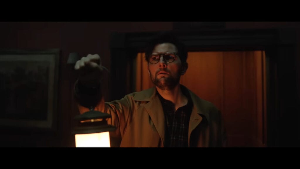 Adam Scott wearing a tan coat and holding up a glowing lantern in a dark hallway.
