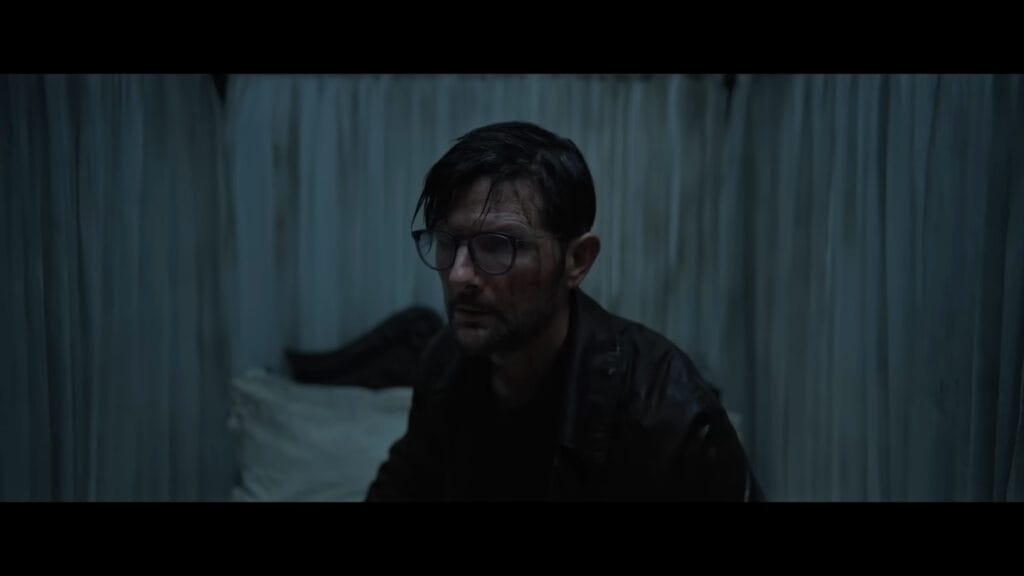 A dishevelled and bruised Adam Scott sitting on a bed surrounded by white curtains in a dark room.