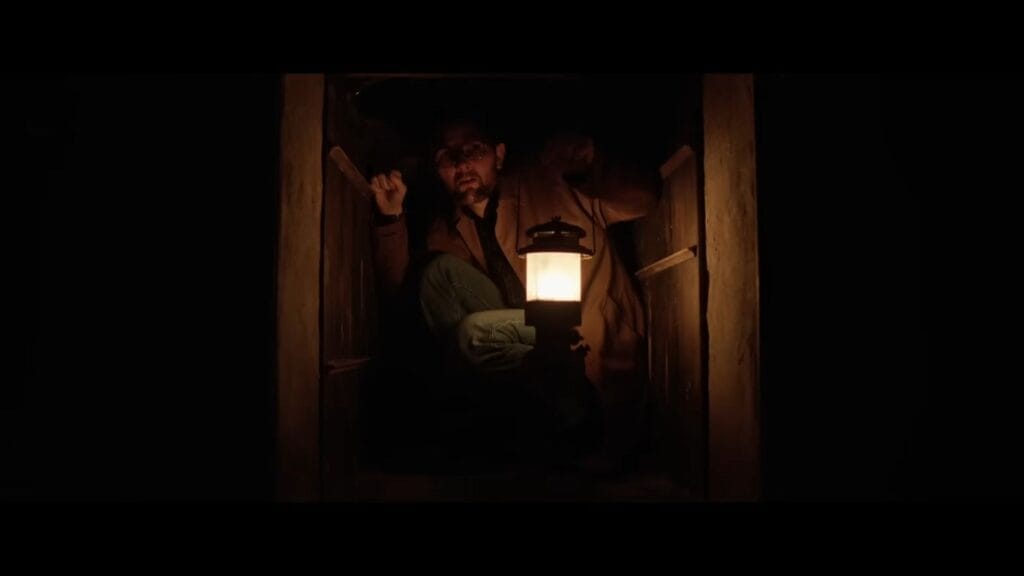 Ohm Bauman (Adam Scott) crouched inside a dark wooden dumbwaiter holding a lit lantern.