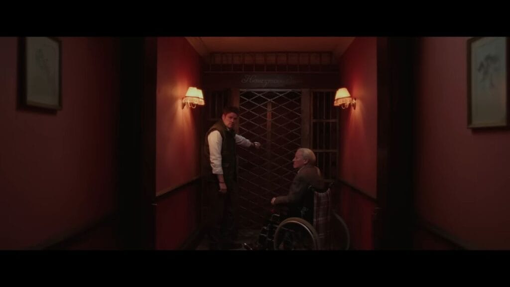 Fergal and Cob in a wheelchair standing in a red hotel hallway before the Honeymoon Suite gate.
