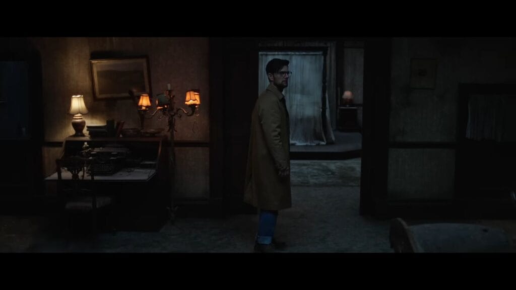 Ohm Bauman standing in a dimly lit, vintage hotel room featuring a typewriter and antique furniture.