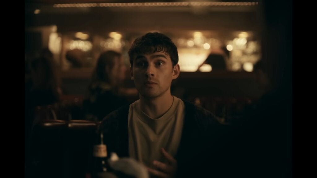 A young man with dark hair looking surprised in a dimly lit bar in the movie Obsession (2025).