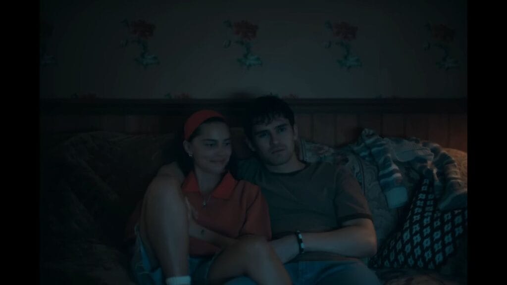 A young man and woman sit together on a sofa in a dimly lit room, watching something off-camera in the movie Obsession (2025)