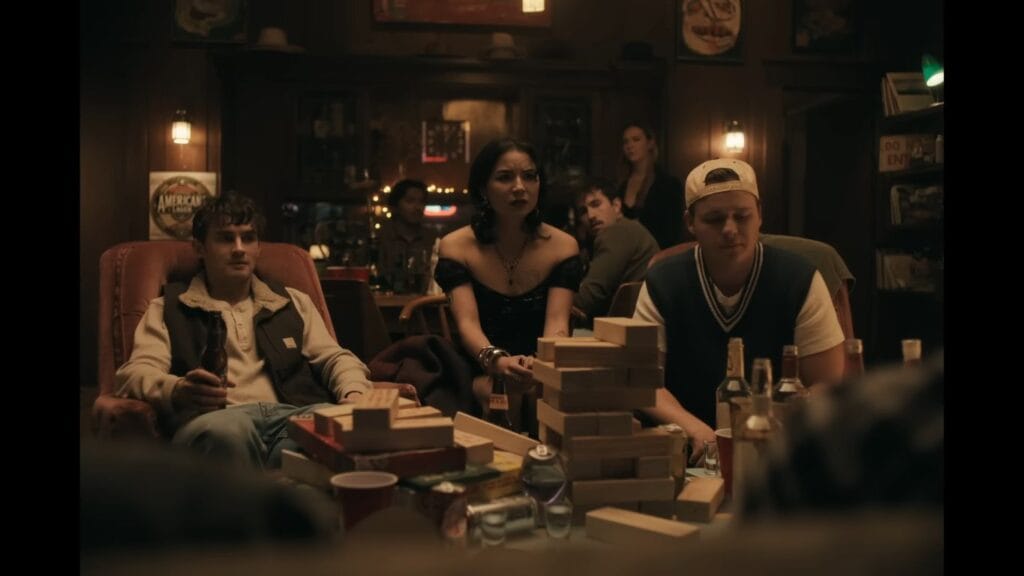 Three young adults sitting in a dimly lit bar around a table with drinks and a wooden block game in the movie Obsession (2025)