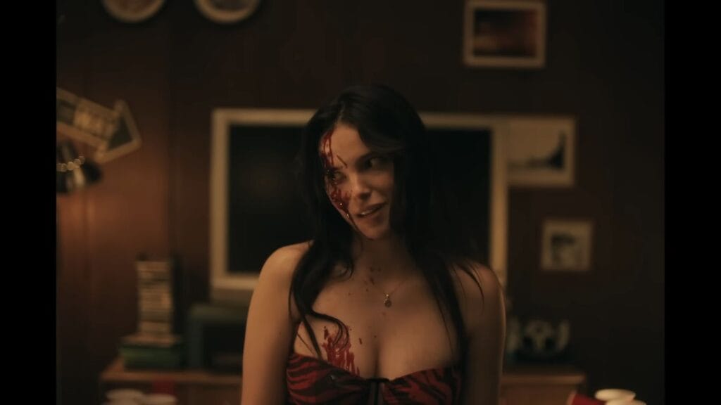 A woman with long dark hair has blood on her face and chest, smiling slightly in a dimly lit room in the movie Obsession (2025)
