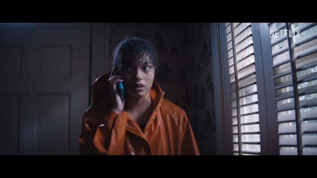 A woman in an orange raincoat looks concerned while talking on a cell phone inside a dimly lit room.