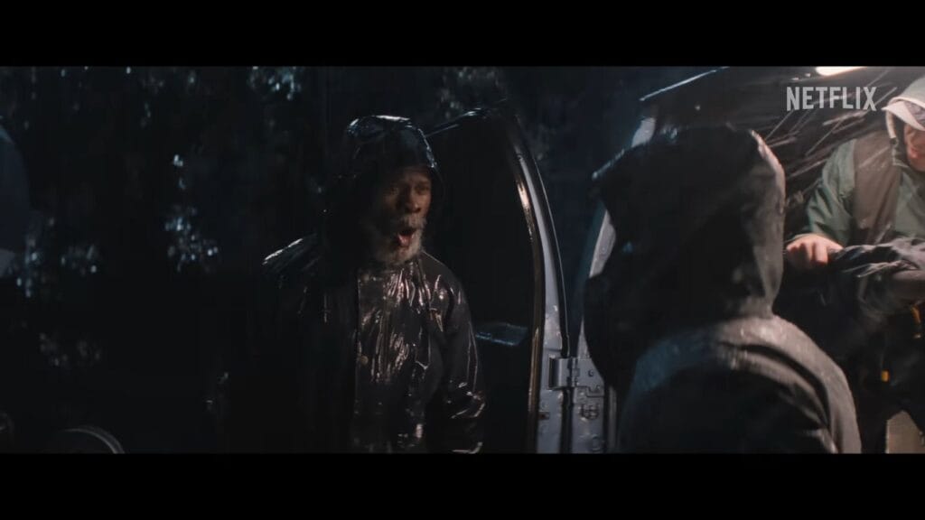 A man in a wet black raincoat shouting toward someone off-camera next to a vehicle at night.