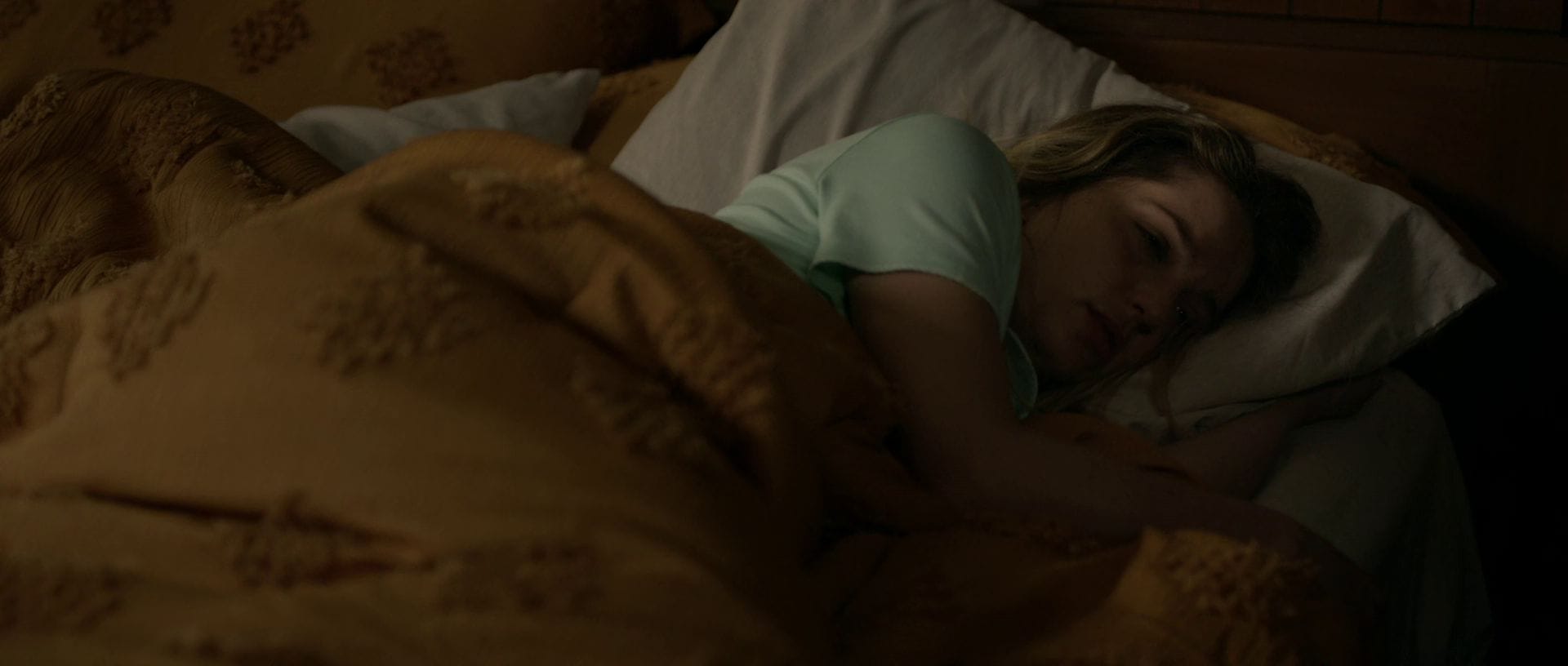 A woman with long blonde hair lies awake and deeply troubled in a dimly lit bedroom under a gold-colored blanket.