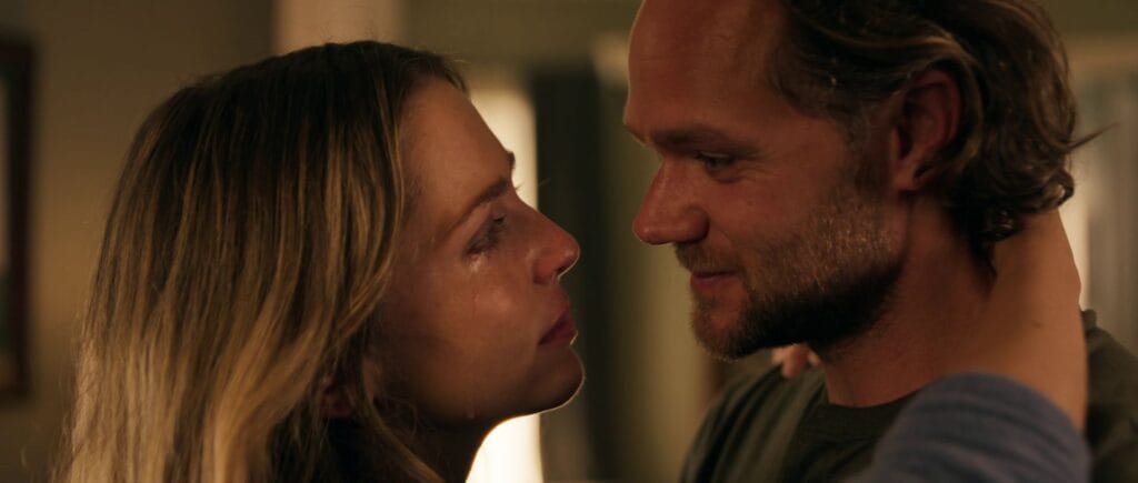 An intimate close-up of a tearful blonde woman locking eyes with a bearded man in a dimly lit room.