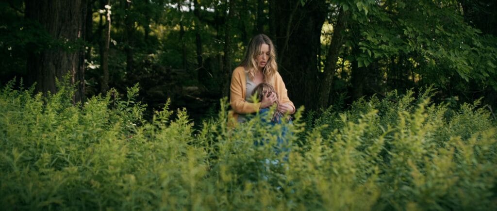 Ellie in a yellow cardigan embraces Alice amidst thick, tall green weeds in front of a dense, dark forest.